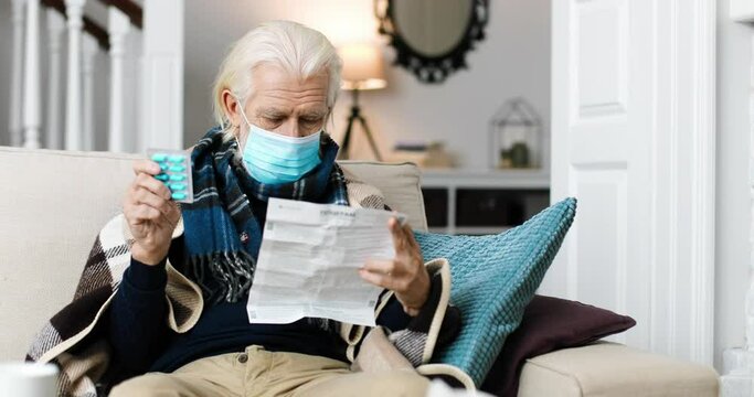 Portrait of grey-haired Caucasian male in mask sitting at home on sofa and reading prospectus for medicine. Sick old male holding blue pills in hand in room. Pandemic virus. Disease treatment concept