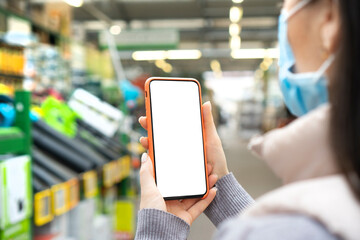 Girl wearing a medical face mask standing in a mall and holding smartphone with empty white screen for copy space.