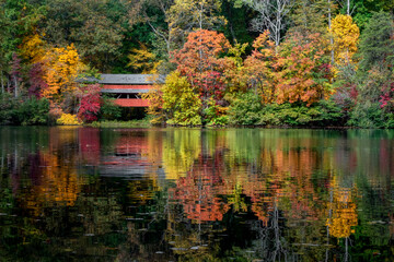 With a beautiful reflection on Lake Loretta in Alley Park, Lancaster, Ohio, the red George Hutchins Covered Bridge, surrounded by colorful autumn leaves, was constructed in 1865 at another location. - obrazy, fototapety, plakaty