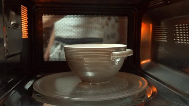 A ceramic bowl with some food is heated in the microwave, spinning , a man's face nervously and discontentedly looks into the microwave through the glass. The man is crazy and hungry.