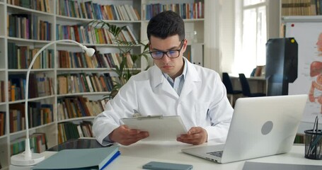 Male doctor reading medical documentation and shaking head while sitting in office. Young man in glasses and white professional gown typing at laptop. Concept of medical worker