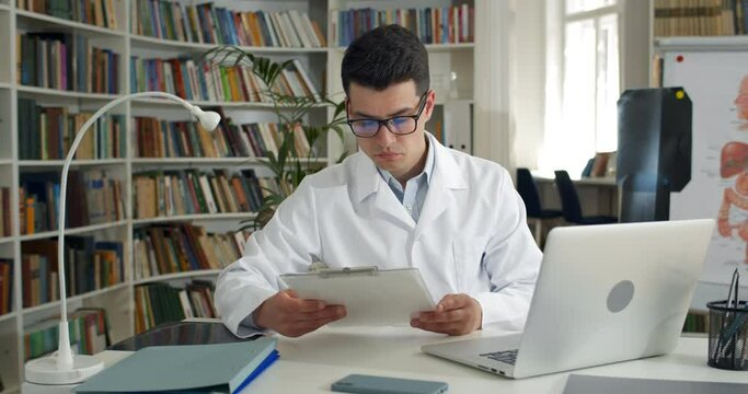Male doctor reading medical documentation and shaking head while sitting in office. Young man in glasses and white professional gown typing at laptop. Concept of medical worker