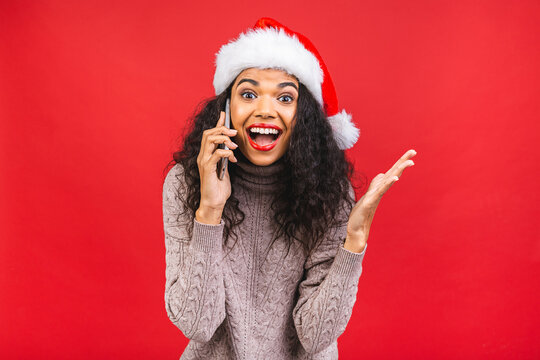 Happy excited african american woman in red santa claus hat with mobile phone isolated over red background.