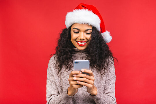 Happy excited african american woman in red santa claus hat with mobile phone isolated over red background.
