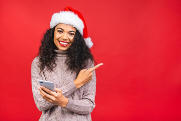 Happy excited african american woman in red santa claus hat with mobile phone isolated over red background. Pointing finger.