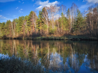 Autumn landscape, forest trees are reflected in calm river water against a background of blue sky and white clouds.