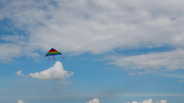 Rainbow kite with tail of three orange ribbons hovers in the blue sky under white Cumulus clouds, while swallows and Swifts fly by quickly.