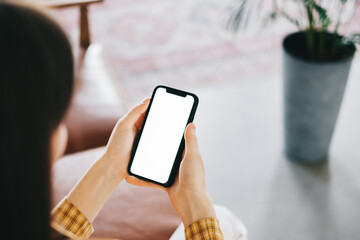 Woman holding a smartphone with a white screen mock up while sitting on the sofa at home.