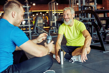 Nice talk after workout. Cheerful middle aged man in sportswear holding bottle of water and talking with fitness instructor while sitting together on the floor at gym