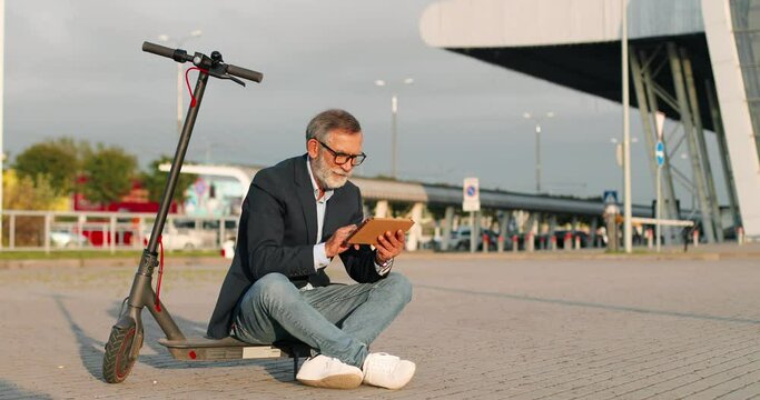 Old Caucasian man in glasses sitting outdoor at electric scooter and using taplet device at station. Male resting and tapping on screen at street. Grandfather browsing online. Businessman pensioner.