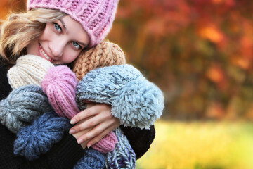 A beautiful young girl holds many knitted hats in her hands. Handmade hats. Woman's portrait on a background of autumn trees. Hobby concept.