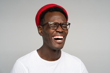 Laughing positive young Afro American man wear red hat in good mood, poses at camera with closed eyes. Overjoyed Black male in spectacles shows her broad smile, isolated on studio grey background. 