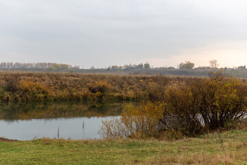 autumn rural natural landscape with calm river water reflecting yellow trees and dry grass in cloudy day