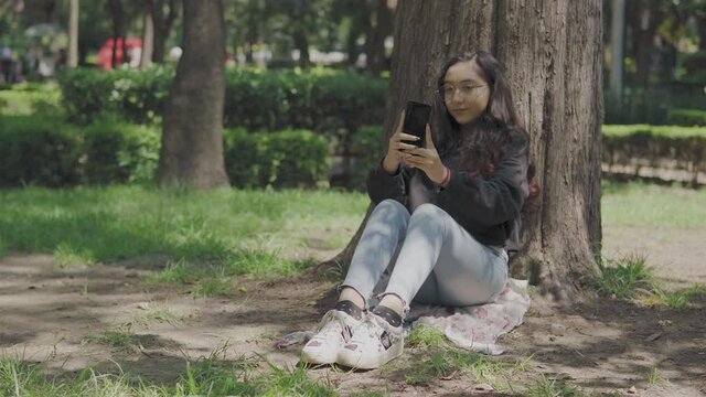 toma horizontal de niña feliz sentada en el pasto en un parque al aire libre tomándose  fotos con su dispositivo móvil, en una tarde soleada 