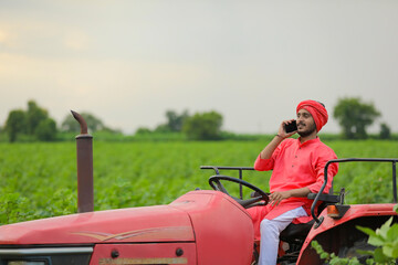 young indian farmer sitting on tractor and talking on smartphone at field