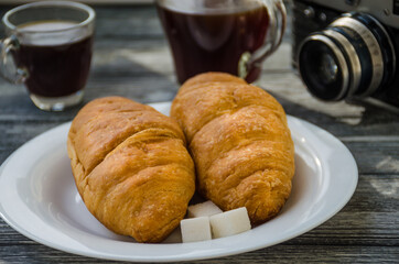 Still life with cup of coffee and croissant on the wooden background. Old retro camera and postcard are near the cup.