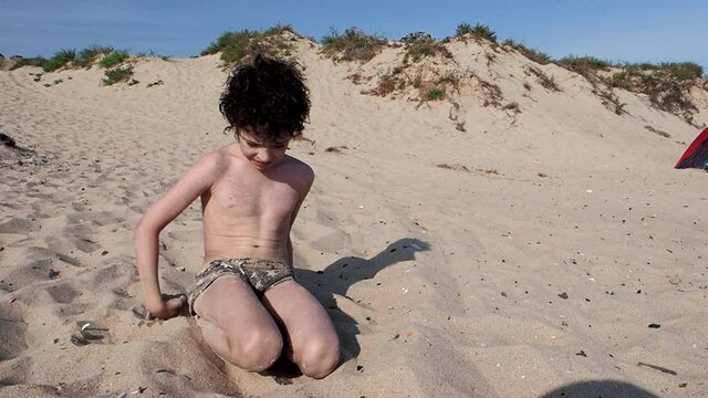 Portait of smiling dark-haired boy playing with sand on the beach and sandy coast background. Child enjoy summer vacation on the seaside. Tourism, resort concept.