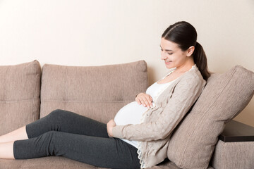 Fototapeta premium photo of cute caucasian future mom lying in light living room on sofa and holding hands on tummy
