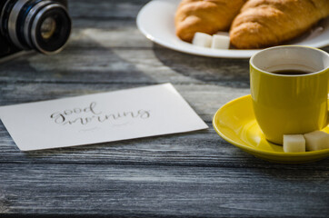 Still life with cup of coffee and croissant on the wooden background. Old retro camera and postcard are near the cup.