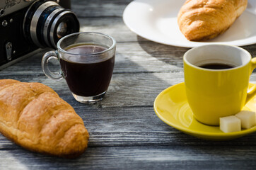 Still life with cup of coffee and croissant on the wooden background. Old retro camera and postcard are near the cup.