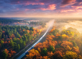 Aerial view of passenger train in beautiful forest in fog at sunset. Autumn landscape with railroad, foggy trees, trail and colorful sky with clouds. Top view of moving train in fall. Railway station