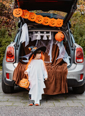 Trick or trunk. Trunk or treat. Little child in Halloween witch hat stands at decorated trunk of...