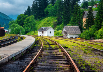 Fototapeta na wymiar Railway station and small white house in mountain village at sunset. Rural railroad in overcast day in summer. Industrial landscape with railway platform, green trees, grass, buildings in cloudy day