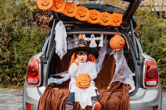 Trick or trunk. Trunk or treat. Little child in witch hat celebrating Halloween party in decorated car trunk eating candies. New trend and alternative safe outdoor celebration of traditional holiday.