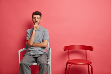 Surprised young Caucasian man keeps hand on chin stares shocked at camera dressed in casual clothes poses near empty chair lives alone poses against rosy background. People home lonely life concept