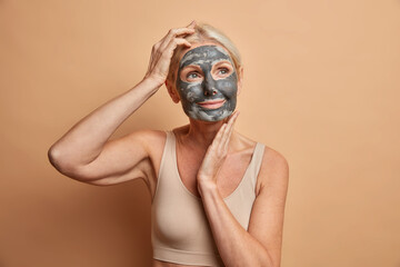 Skin care concept. Dreamy thoughtful senior blonde woman applies moisturizing mask for reducing...
