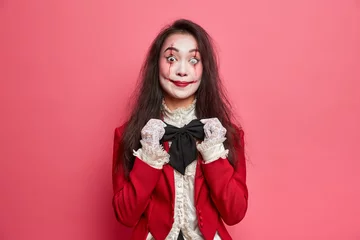 Tuinposter Mysterious brunette woman vampire adjustes black bowtie has scary makeup and bloody scars prepares for halloween isolated over pink background. Spooky zombie wears contact lenses ready for party © Wayhome Studio