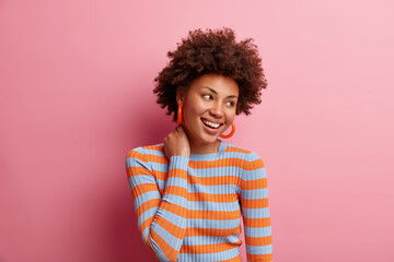 Photo of curly haired cheerful millennial girl touches neck and looks happily away dressed in casual striped jumper being in good mood isolated on pink studio background. Human emotions concept