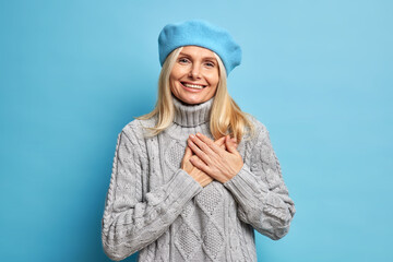 Smiling beautiful woman keeps hands pressed to heart expresses gratitude wears blue beret and...