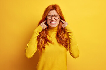 Studio shot of dissatisfied redhead woman clenches teeth and plugs ears annoyed by loud sound or noise wears casual jumper isolated on yellow background. Female complains on music from outside