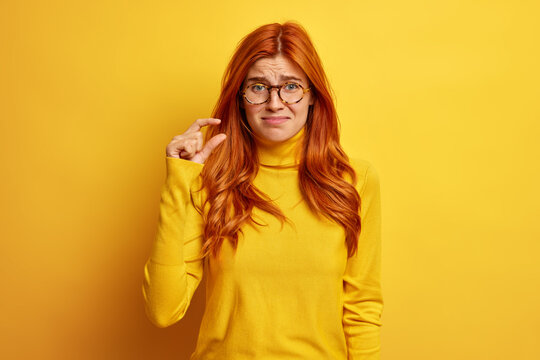 Horizontal shot of unhappy redhead woman makes little gesture demonstrates something tiny dressed in casual jumper poses indoor. Dissatisfied female student shows ammount of work she finished