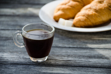 Still life with cup of coffee and croissant on the wooden background. Old retro camera and postcard are near the cup.