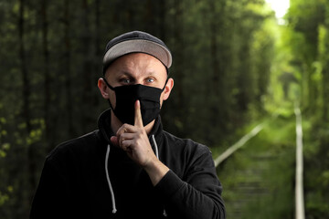 Young man in black protective antiviral mask with a finger to his lips in the summer park. The guy is resting outdoors in a mask made by his own hands. Virus protection. Quarantine measures