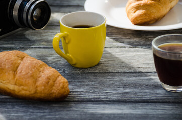 Still life with cup of coffee and croissant on the wooden background. Old retro camera and postcard are near the cup.