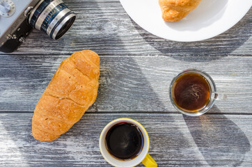 Still life with cup of coffee and croissant on the wooden background. Old retro camera and postcard are near the cup.