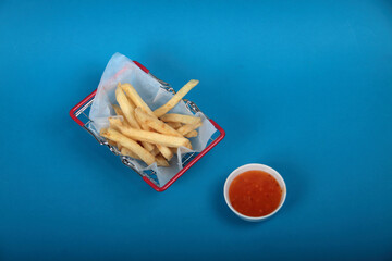 shopping basket of french fries on parchment paper and a bowl of sauce on blue background flat lay