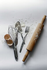 Kitchen utensils in flour on a white background. Old stainless spoons, fork. A whisk and a rolling pin. Egg shell. A broken egg. View from above.