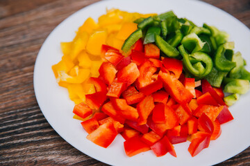 Red, yellow and green bell peppers, diced on a white plate. Dish on a wooden dark background