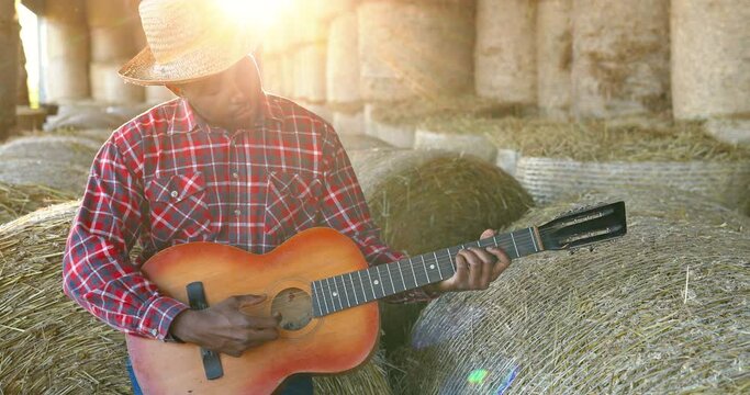 African American young handsome man sitting on hay stocks in shed and playing music on guitar. Male farmer singing and play on musical instrument song. Countryside concept. Village. - Powered by Adobe