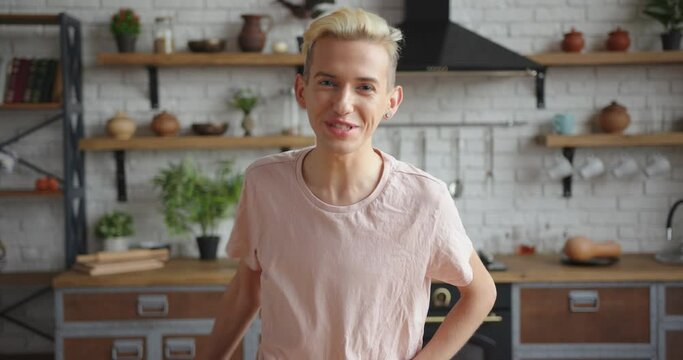 Joyful young man with blond colored hair in pink casual t-shirt talks looking into camera in contemporary kitchen at home