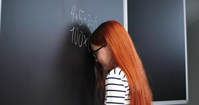 Caucasian red-haired girl standing at blackboard and writing hard equations. Schooling concept. Schoolgirl doing difficult math at lesson in school and leaning with forehead on board. Mathematics.