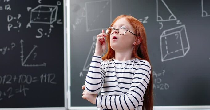 Portrait. Small cute clever girl in glasses and with red hair standing in classroom and thinking about mathematics. Little nice smart schoolgirl finding out idea and right decision in math. Blackboard