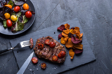 Roasted beef sirloin steak served with vegetable crisps chips, salad and wine on black slate board