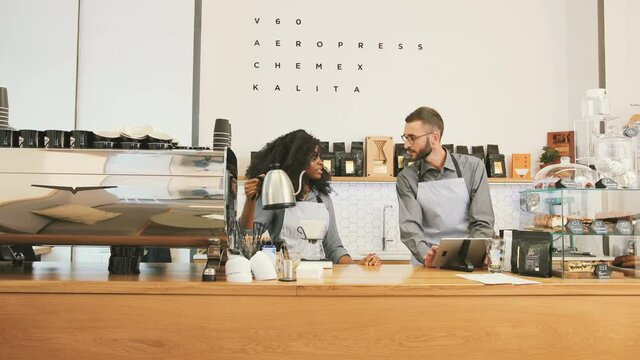 Beautiful African-American barista brewing coffee at bar. Two cafe employees talking while working. Barista consults colleague and uses tablet.