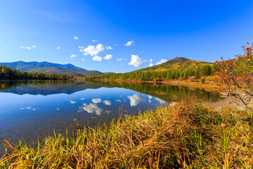 Naklejka premium The nature of the Magadan region. A beautiful flat surface of the lake against the background of colored hills and the blue sky. Fascinating view of the forest lake