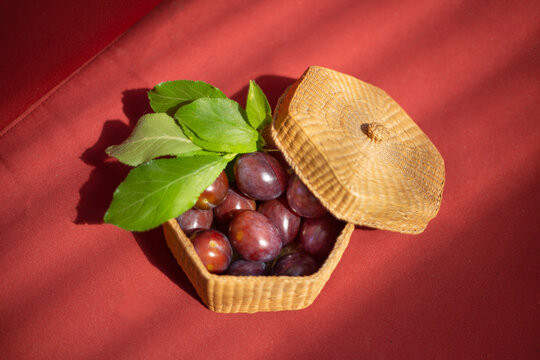 An isolated and sunlight dappled view of black plums with green leaves in a lidded basket against a rust colored background

